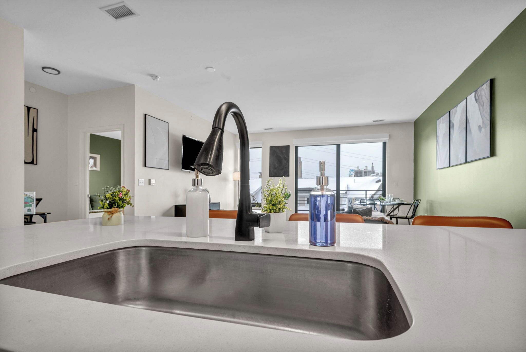 A modern kitchen with a stainless steel sink, a black faucet, and a white countertop. There are two potted plants on the counter.