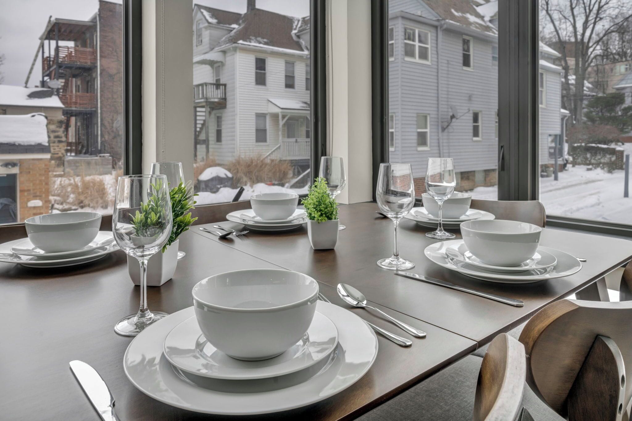 Mesa de jantar posta com tigelas, pratos e copos brancos, com vista para casas através de grandes janelas.