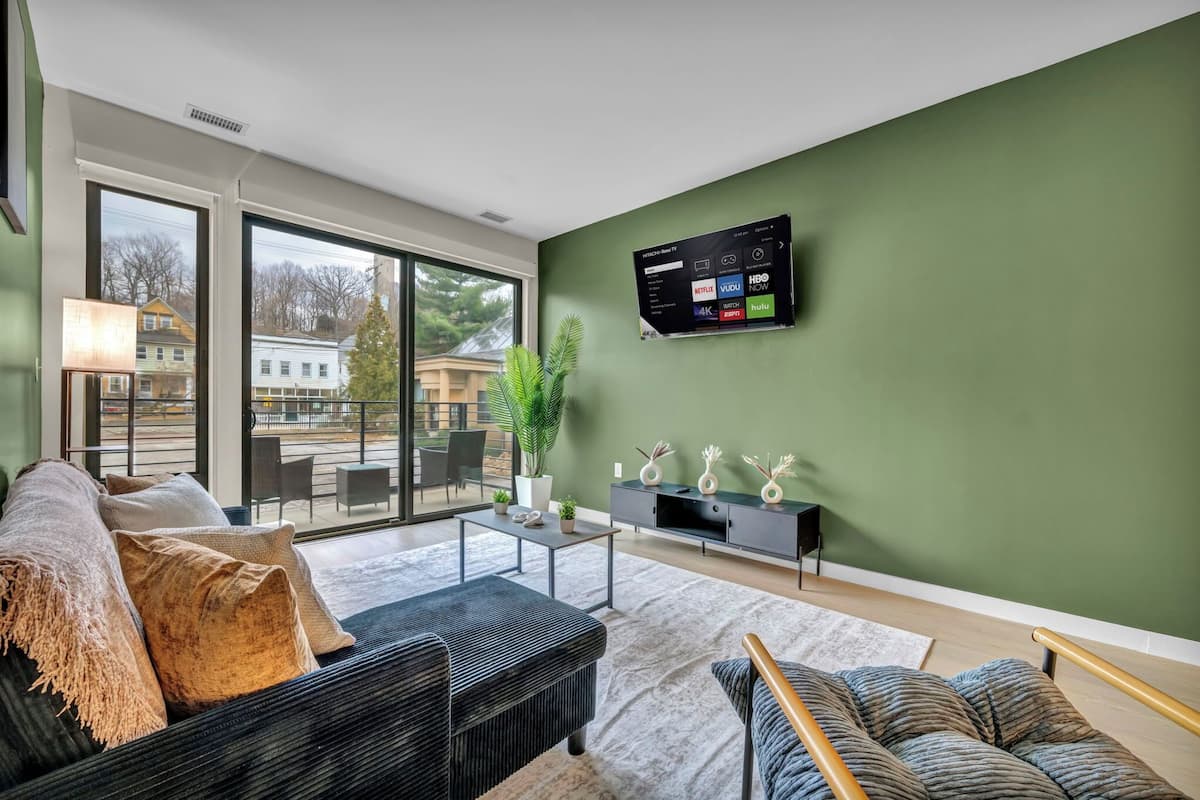 A modern living room with a large TV, comfortable seating, a plant, and a view through large windows.