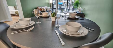 A dining table set with white bowls and wine glasses, surrounded by black chairs, in a room with green walls and a view of buildings outside.