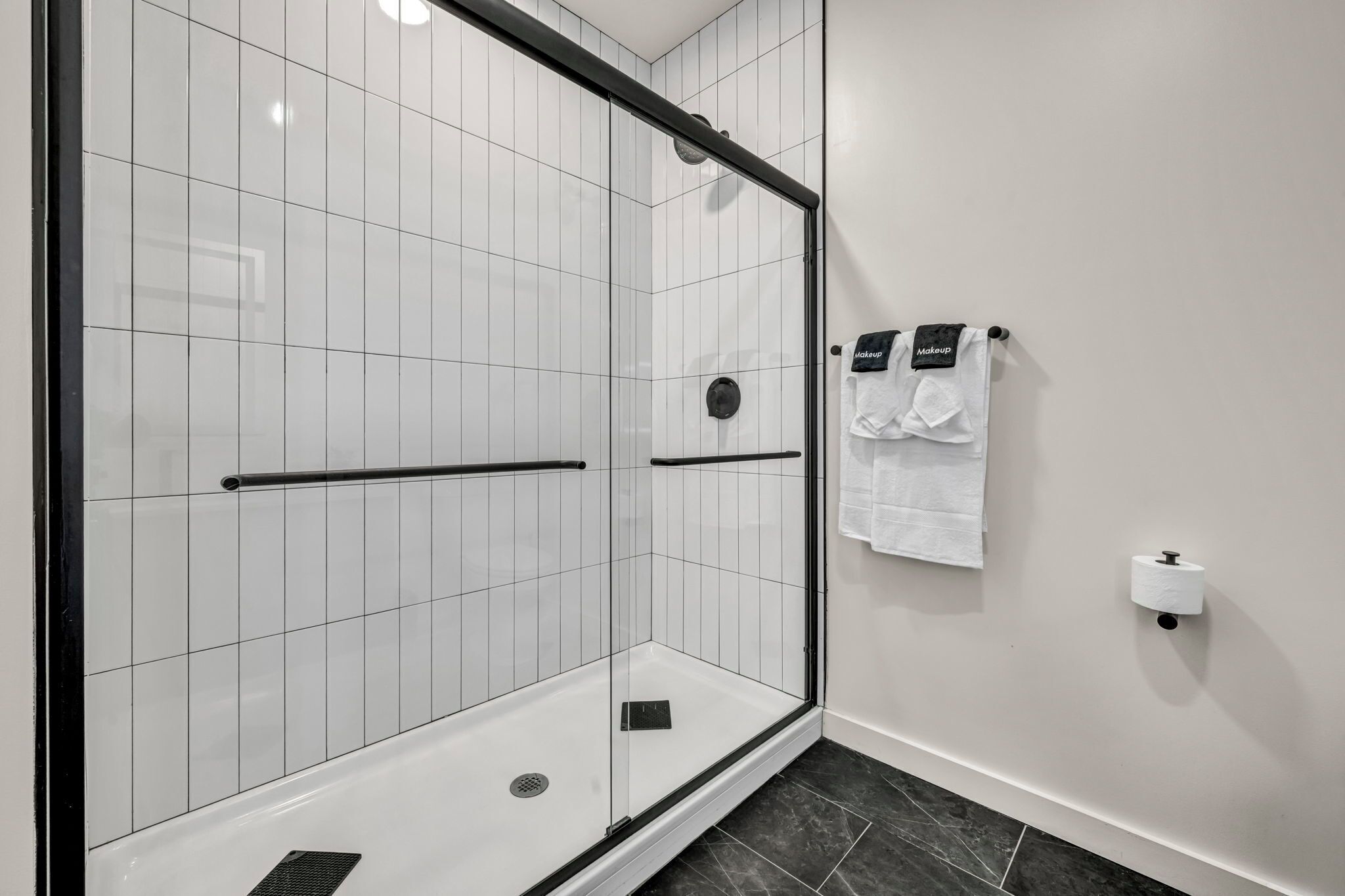 A modern bathroom with a glass-enclosed shower, white tiled walls, a black towel rack, and a toilet paper holder.