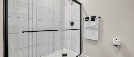 A modern bathroom with a glass-enclosed shower, white tiled walls, a black towel rack, and a toilet paper holder.