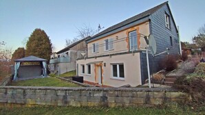 A two-story house with a balcony, a small attached garage, and a stone retaining wall. - Cosy holiday flat in a quiet location, close to Luxembourg and Trier (Ralingen)