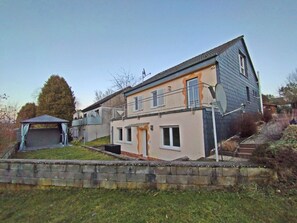 Ein zweistöckiges Haus mit Balkon, einer kleinen angebauten Garage und einer Steinmauer.