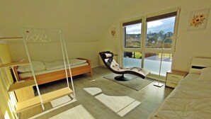 A bedroom with a bunk bed, a reclining chair, a bedside table, and a large window with a view of houses. - Cosy holiday flat in a quiet location, close to Luxembourg and Trier (Ralingen)