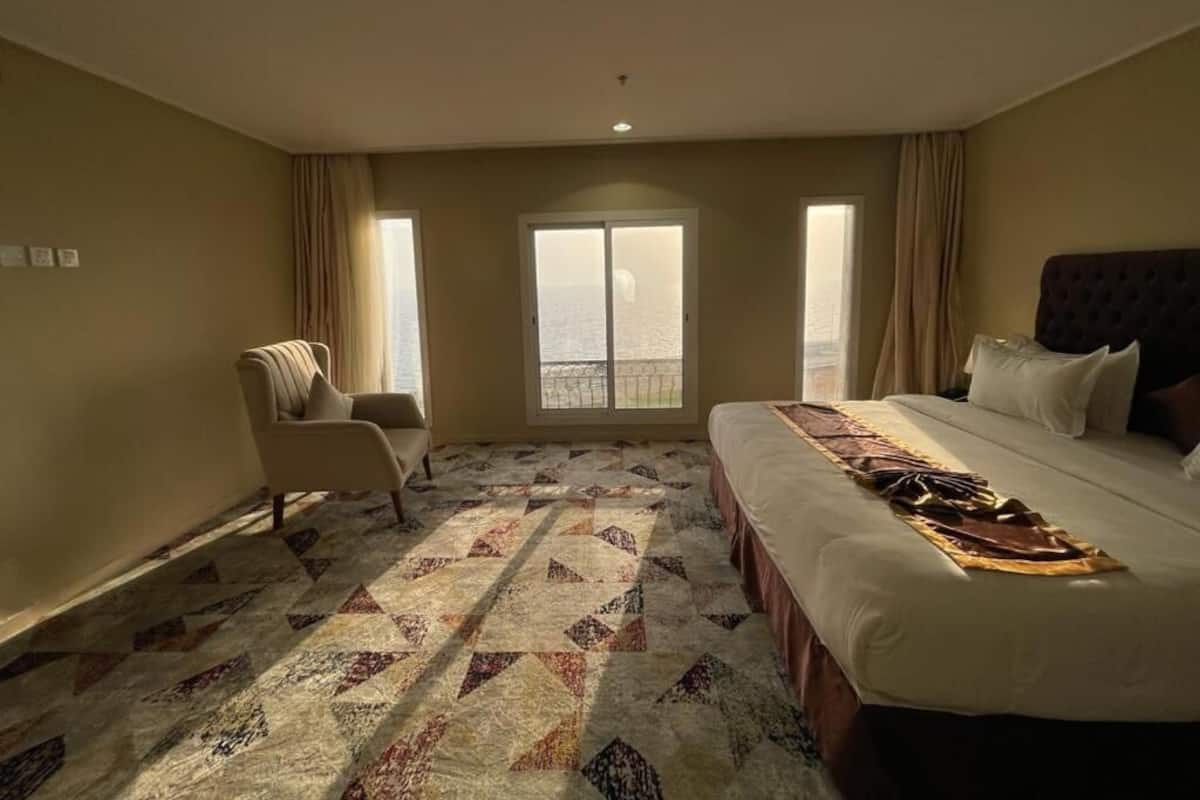 A hotel room with a large bed, a chair, a patterned carpet, and a view through a window.