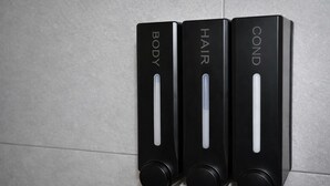 Trois distributeurs noirs fixés au mur, portant les inscriptions « CORPS », « CHEVEUX » et « APRÈS-SHAMPOING », sont disposés sur un mur carrelé.
