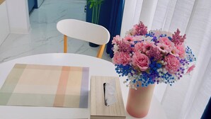 Una habitación con una mesa redonda, una caja de pañuelos, un florero con flores, una silla y una lavadora al fondo.