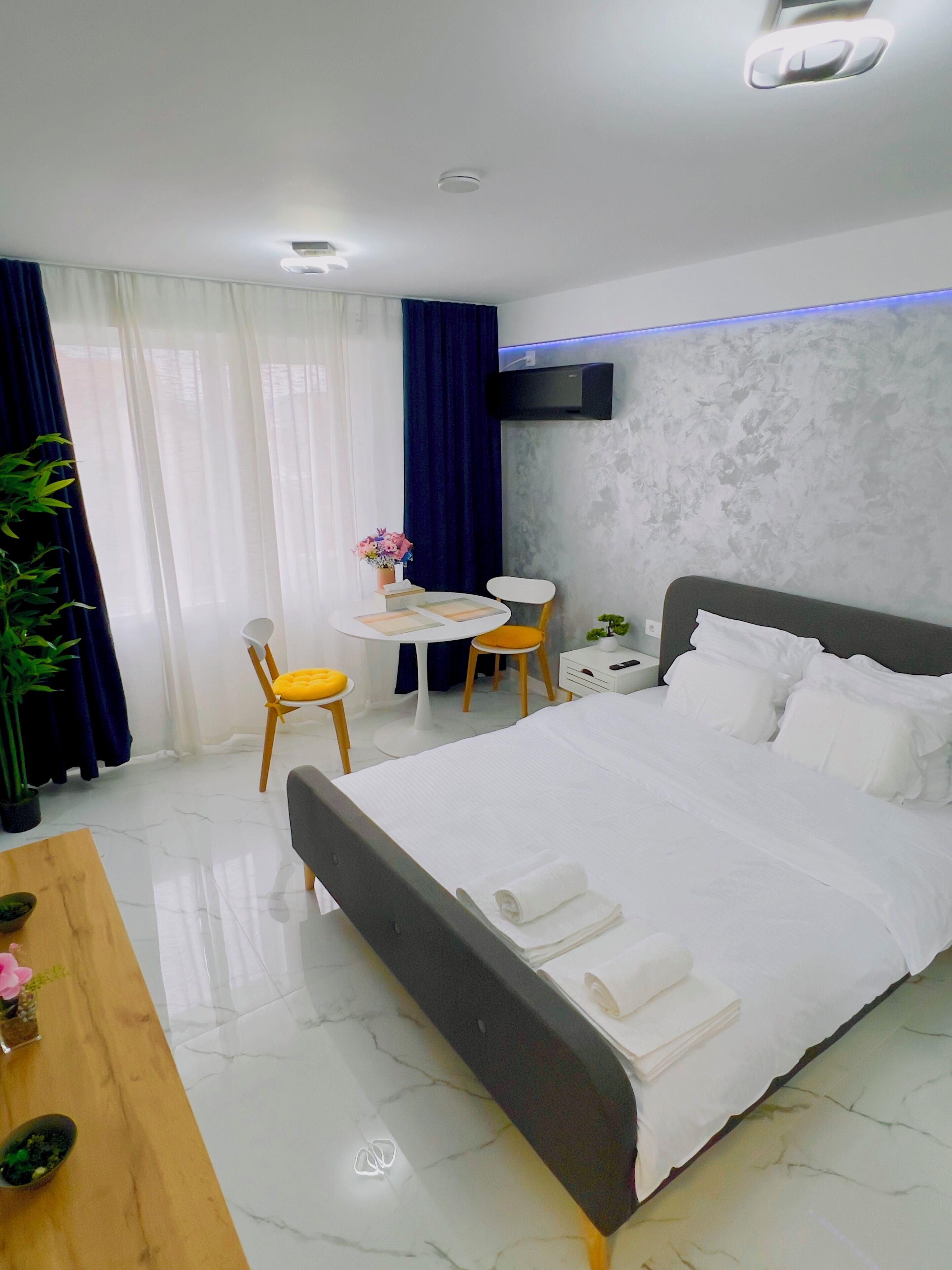 A modern hotel room with a large bed, a desk with a chair, a small table with a chair, and a potted plant.