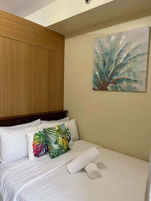 Ein Hotelzimmer mit Bett, weißen Leinentextilien, einem Holz-Kopfteil, einem tropisch gestalteten Kopfkissen und einem Pflanzenbild an der Wand.