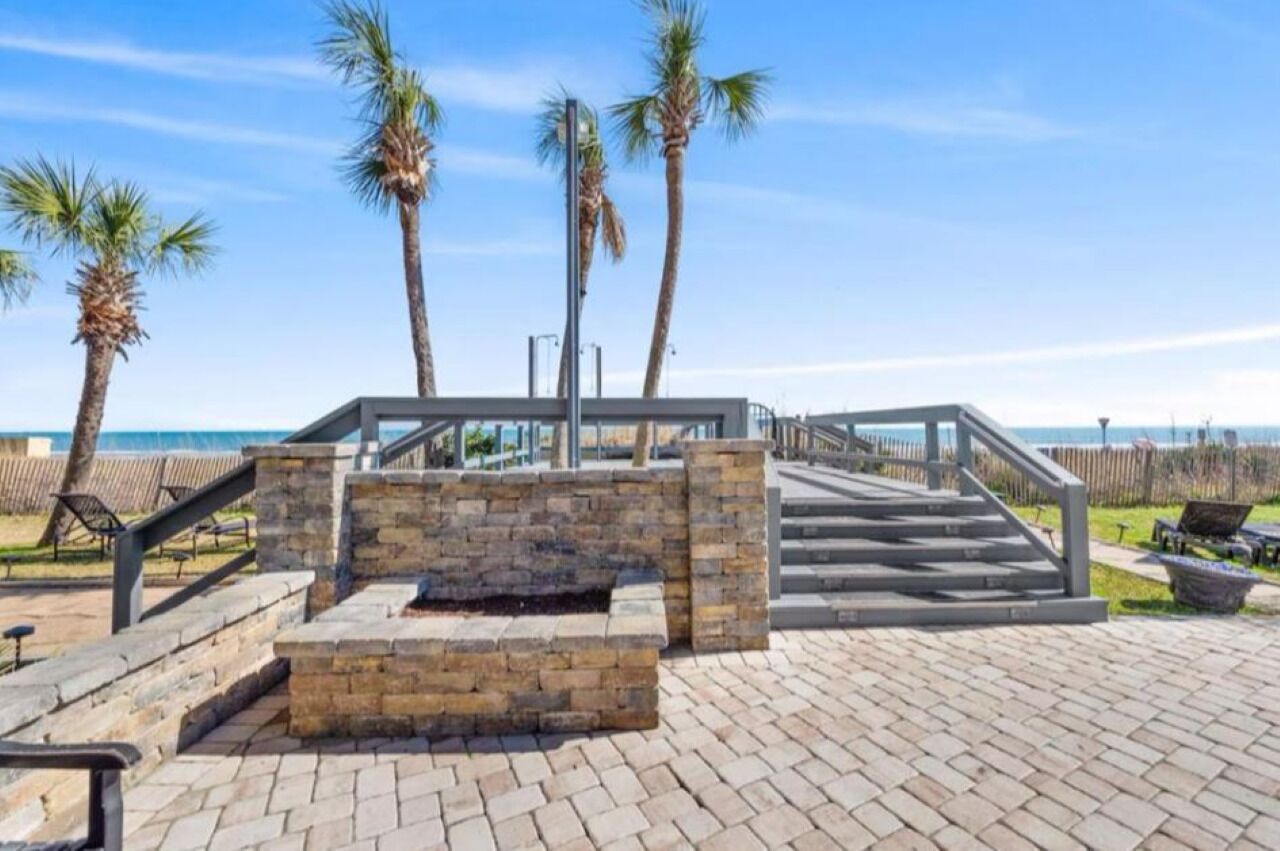 A paved walkway with stone steps leading to a seating area with a fire feature, palm trees, and a view of the beach.