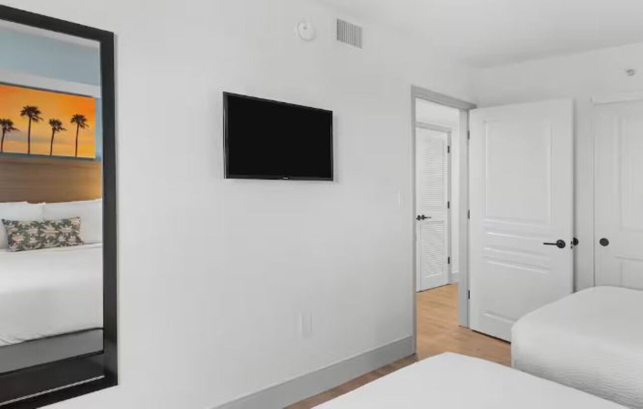 A hotel room with a bed, a flat-screen TV mounted on the wall, a door, and a mirror reflecting part of the room.