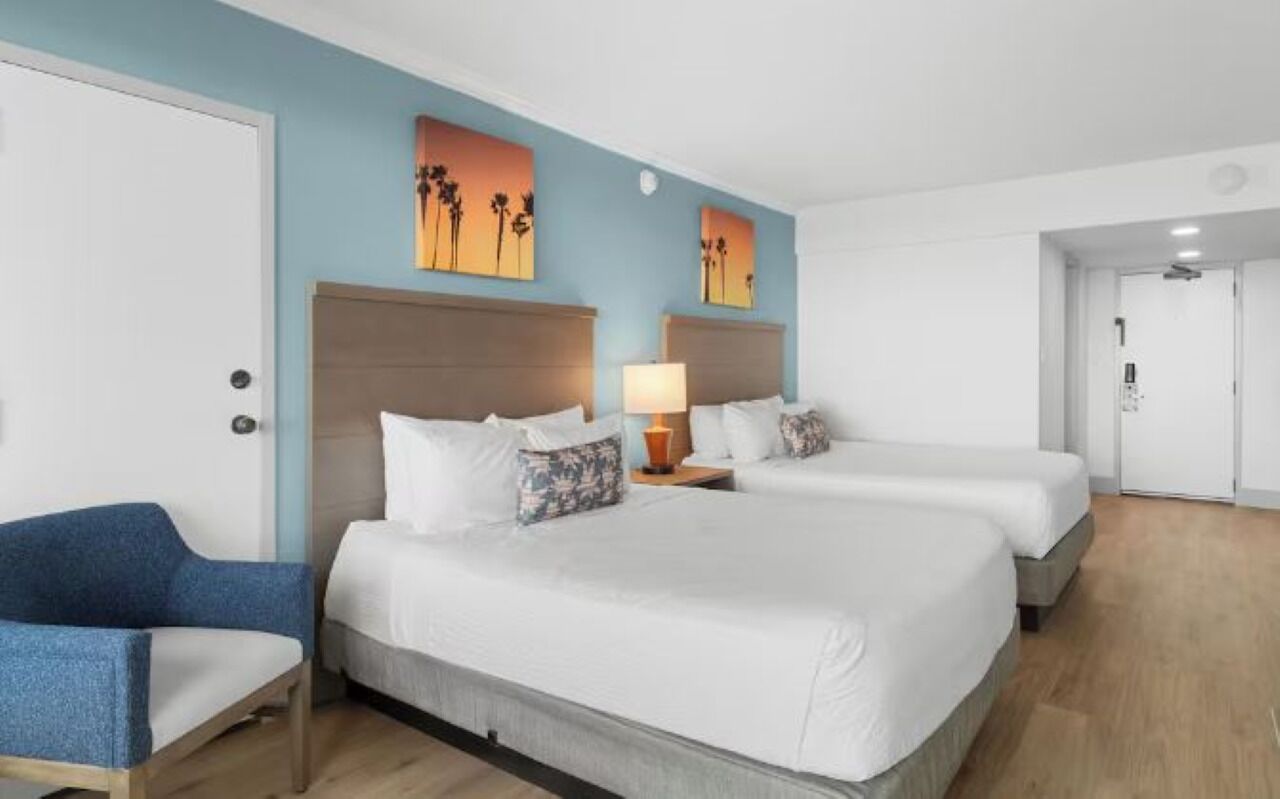 Une chambre d’hôtel avec deux lits, un fauteuil bleu et des œuvres d’art accrochées aux murs.