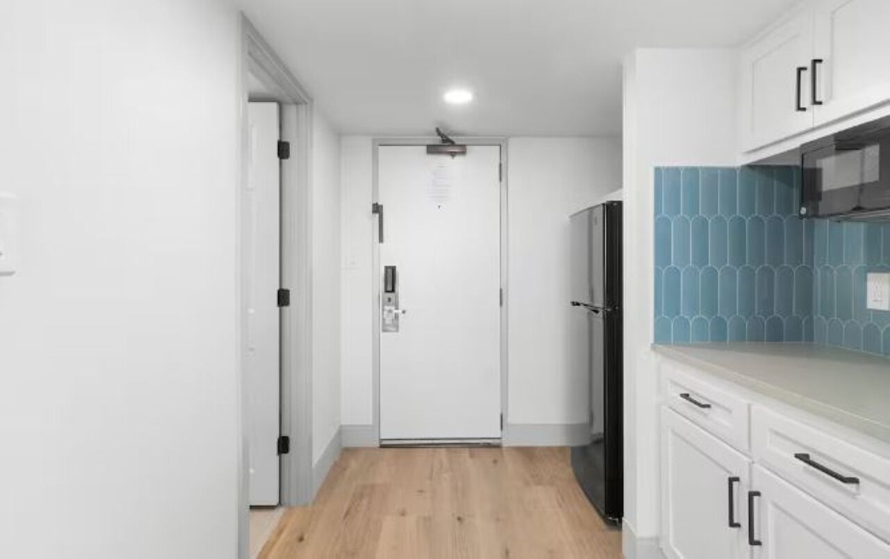 Un pasillo estrecho con una puerta blanca al final, un refrigerador negro y armarios blancos con manijas negras.