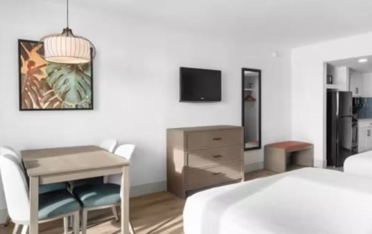 A modern hotel room with a bed, a desk with chairs, a dresser, a television, and a painting on the wall.