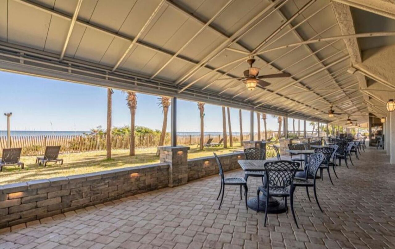 Un patio exterior con suelo de ladrillo, sillas y mesas de metal, con vista a una playa con palmeras y una cerca.
