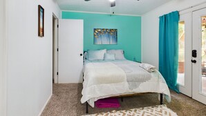 Un dormitorio con cama, ventilador de techo, un cuadro, una puerta y una ventana con cortinas.