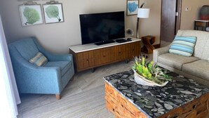 Un salon équipé d’un téléviseur à écran plat, d’un fauteuil bleu, d’un canapé beige, d’une table basse agrémentée d’une plante et d’un meuble en bois.