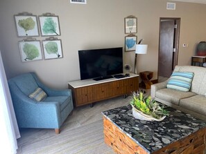 Ein Wohnzimmer mit Flachbildfernseher, einem blauen Sessel, einem beigen Sofa, einem Couchtisch mit Pflanze und einer Holzkommode.