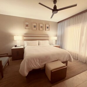 Una habitación de hotel con una cama grande, dos mesitas de noche con lámparas, un ventilador de techo y un ventanal con cortinas traslúcidas.