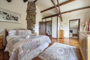 A bedroom with a large bed, a stone wall, wooden beams, a wooden dresser, and a mirror. - 16th Century Devon Farmhouse, Stunning Views (Winkleigh)
