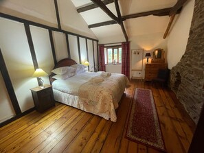 A bedroom with a wooden floor, a bed with white bedding, a nightstand with a lamp, a dresser, and a window with curtains. - 16th Century Devon Longhouse, Stunning Views (Winkleigh)