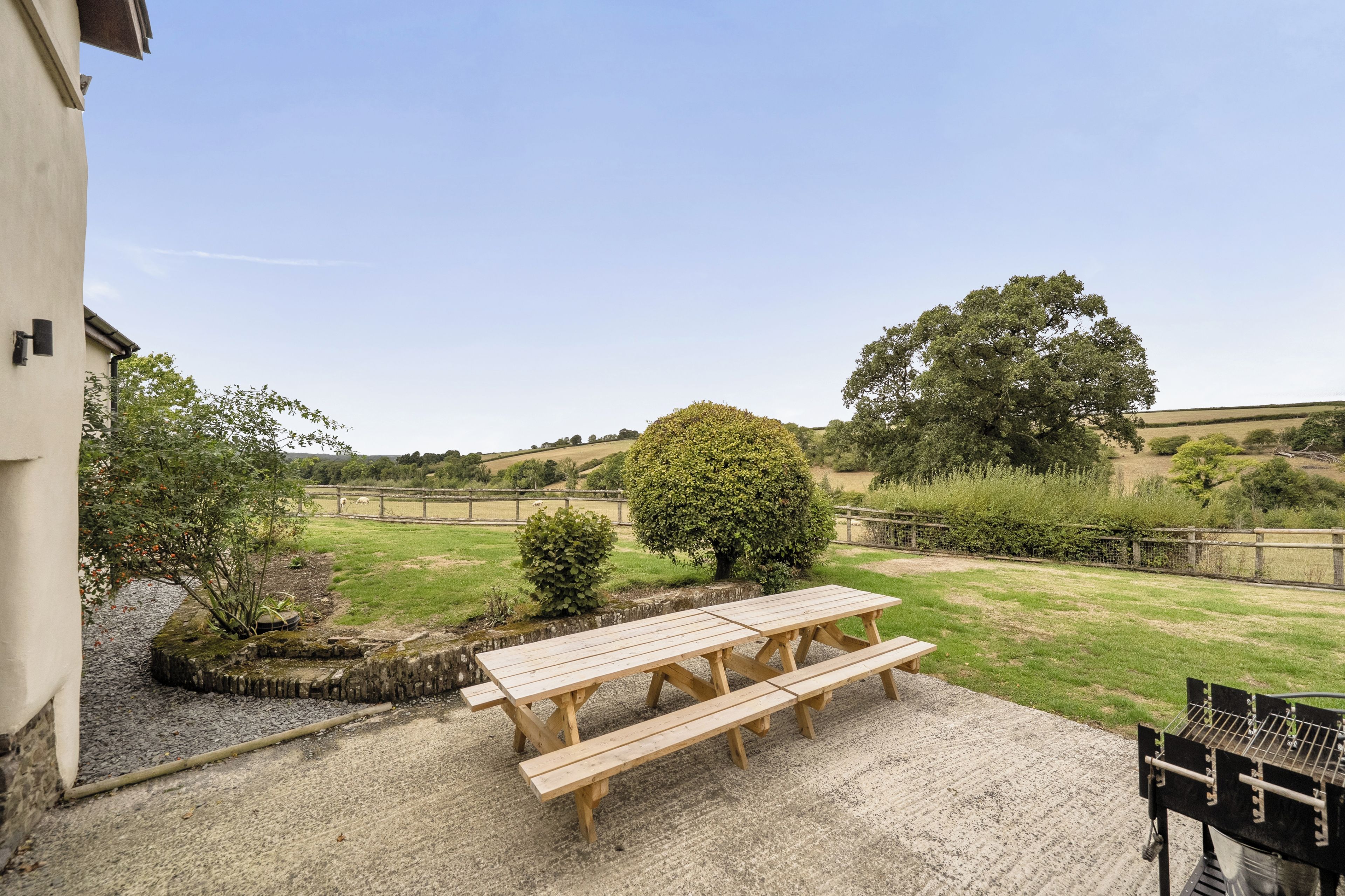 Un patio con una mesa y bancos de madera, rodeado de grava y un jardín cercado con árboles y vistas a colinas ondulantes.