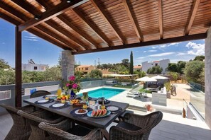 An outdoor dining area with a pool view, wicker furniture, and a clear sky. - Villa Ampelaki (Chania)