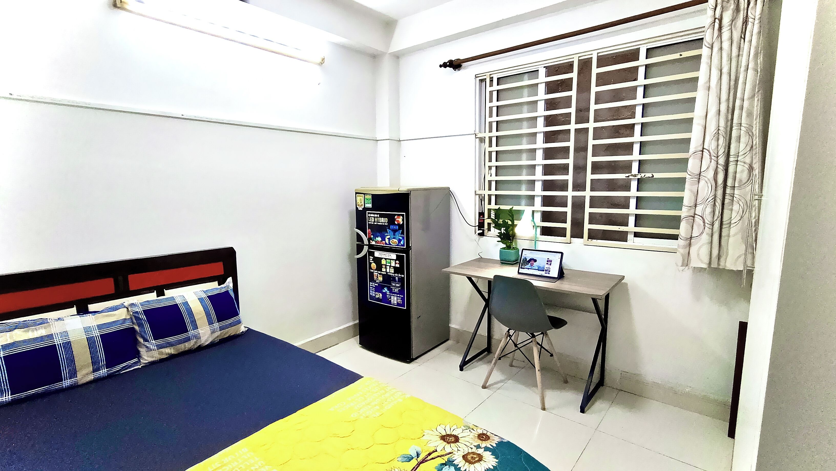 A small, clean room with a bed, a desk, a refrigerator, and a window with blinds.