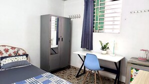 Un dormitorio con cama, escritorio, silla, armario, ventana con cortinas y una mesita con planta y tetera.