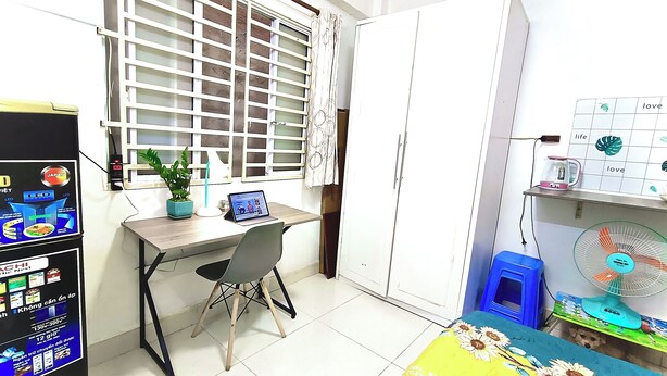 Habitación compacta con escritorio, silla, planta en maceta y un pequeño ventilador.