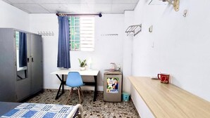 Una habitación pequeña y ordenada con cama, escritorio, silla, armario, ventana con cortinas, una mesita con una taza y un dispensador de agua.