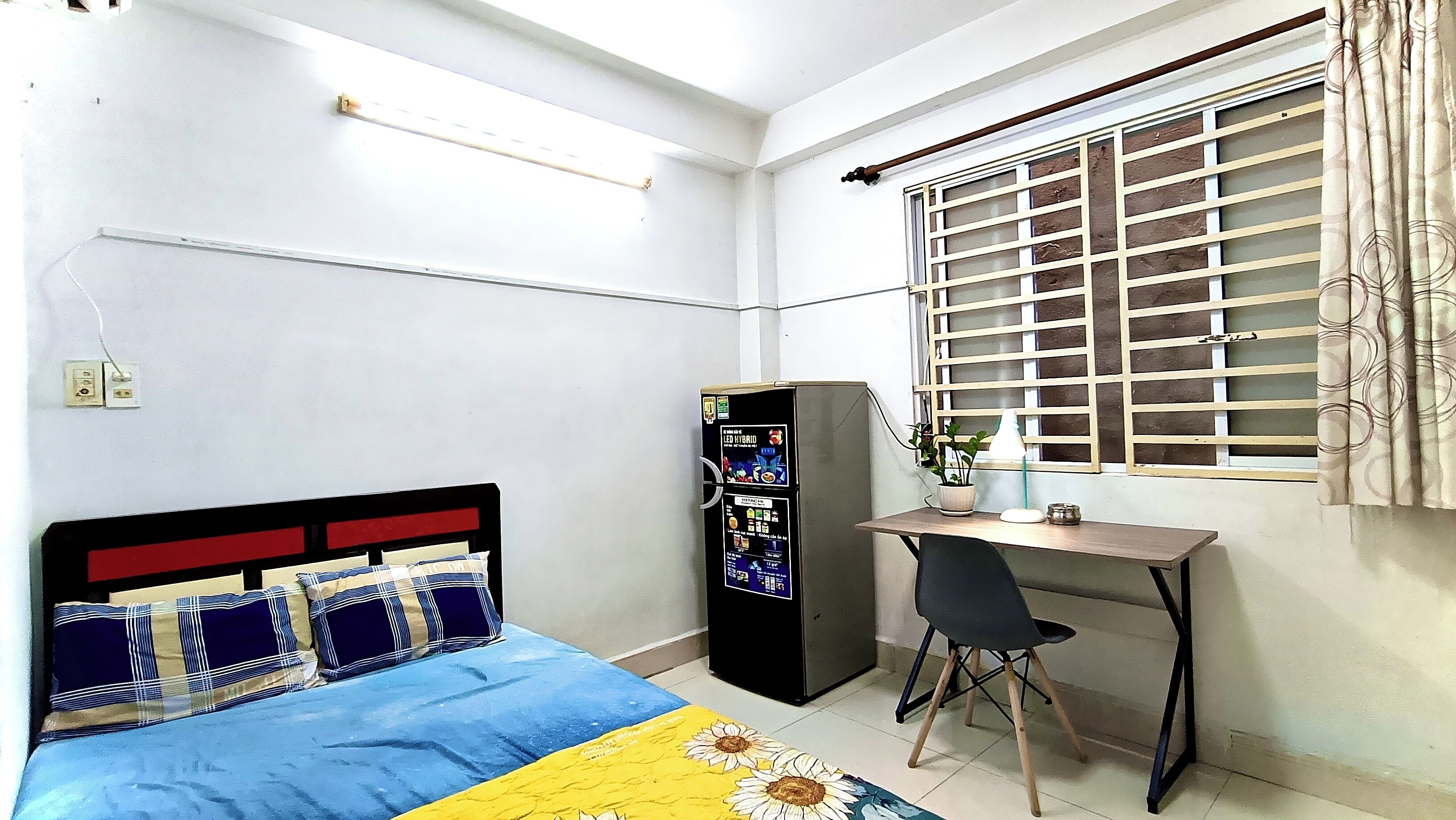 A bedroom with a bed, desk, chair, refrigerator, and window with blinds.