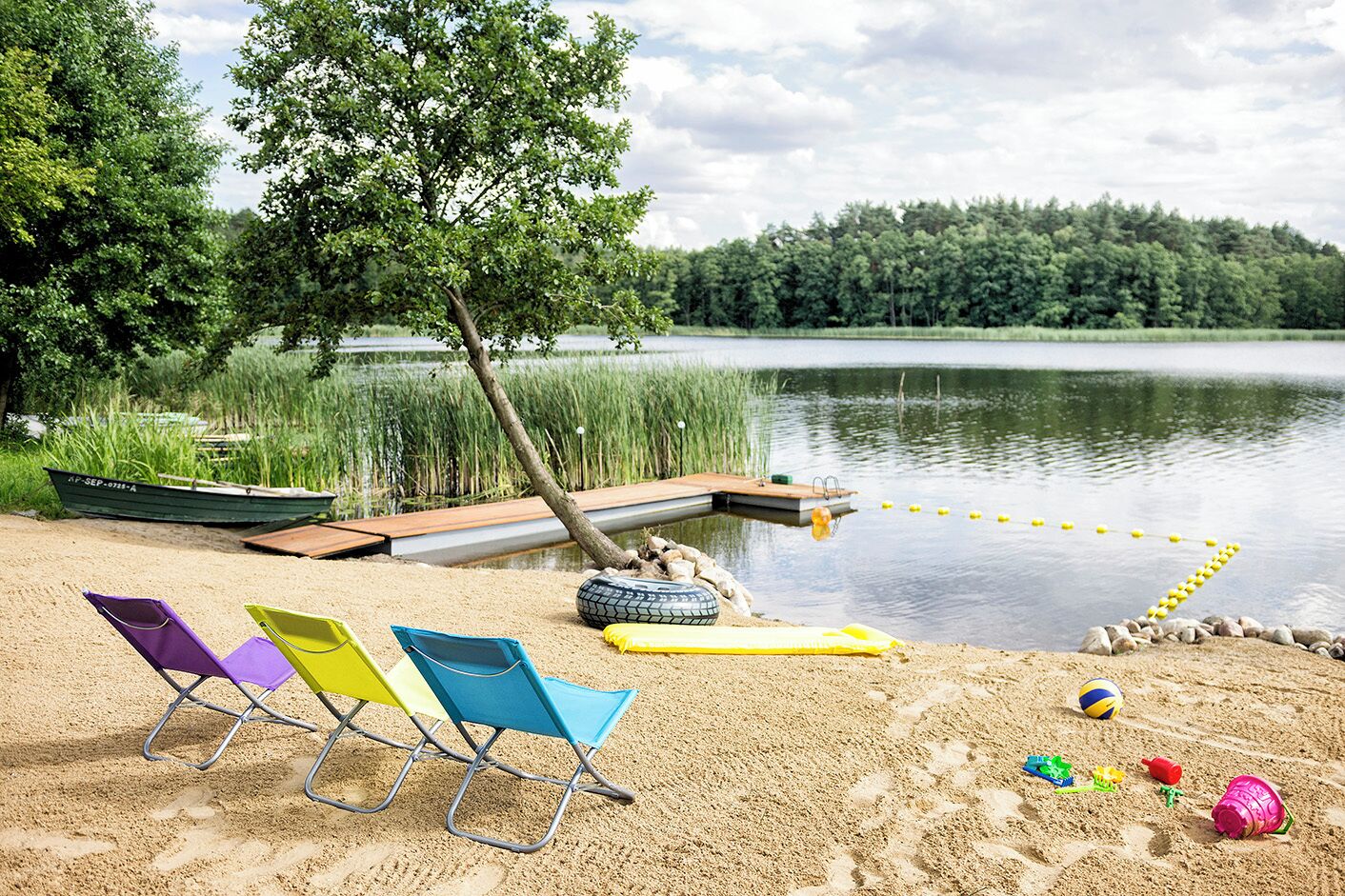 A lakeside beach with a boat, colorful beach chairs, a tire raft, and a volleyball.