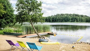 Une plage au bord d’un lac, avec un bateau, des chaises de plage colorées, un radeau en forme de pneu et un filet de volley-ball.