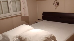 Un letto rifatto con lenzuola e cuscini bianchi, una testata scura, una finestra con persiane bianche e uno scaffale a parete con un piccolo oggetto.