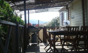 Un porxo cobert amb taula i cadires, un ventilador i vista a una casa i muntanyes.