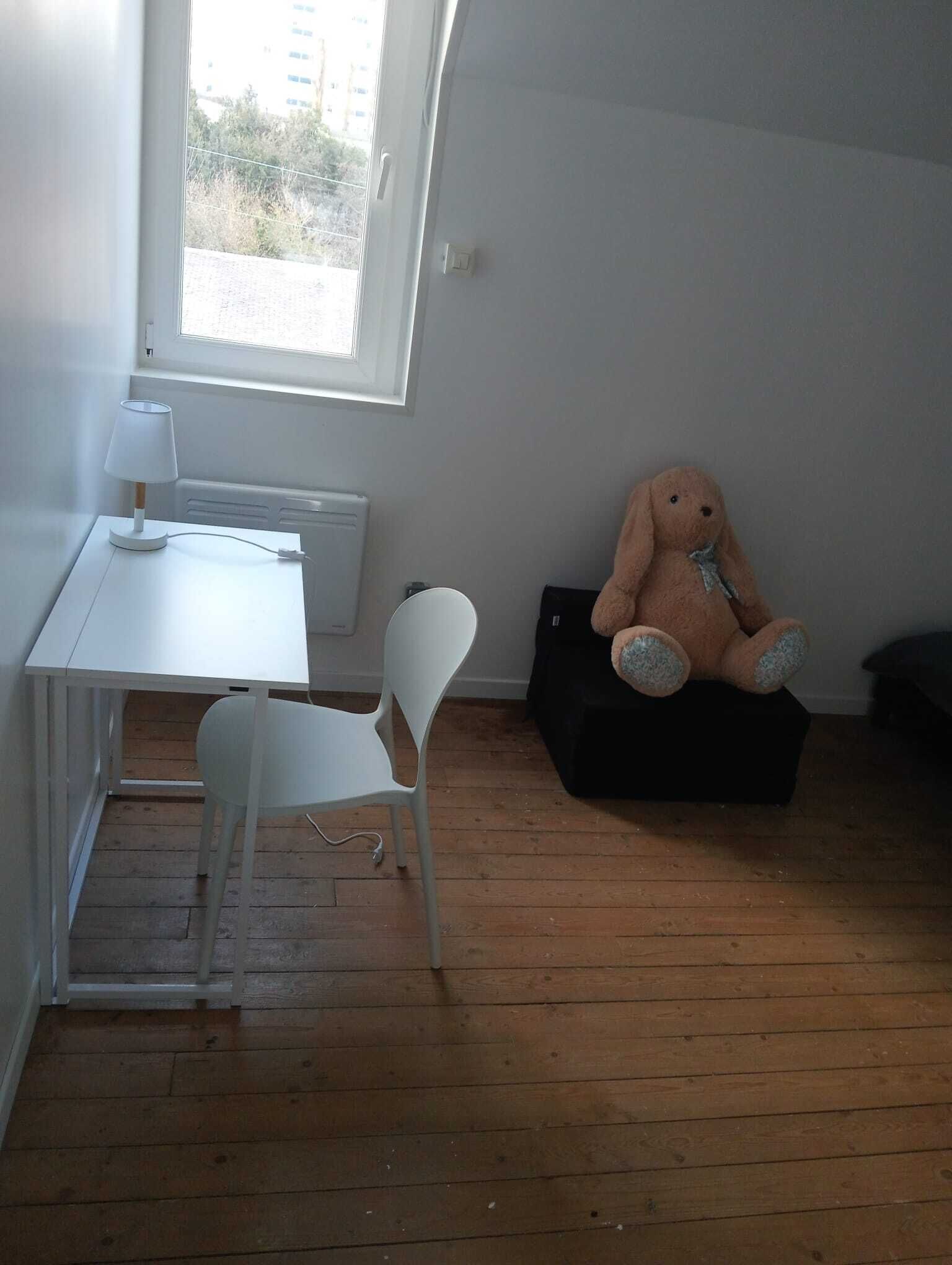 Une petite pièce avec un bureau blanc, une chaise blanche, une grande fenêtre et un jouet en peluche posé sur un pouf noir.