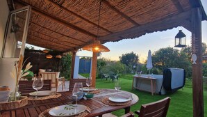 Un espace de restauration en plein air avec une table en bois dressée pour deux personnes, entourée de chaises, et un toit de chaume au-dessus.
