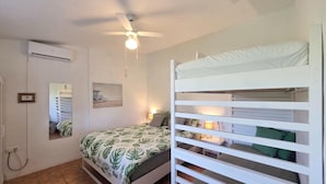 Une chambre avec un lit superposé, un ventilateur de plafond, un climatiseur fixé au mur, une lampe de chevet et un tableau encadré accroché au mur.