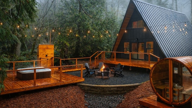 Eine Holzhütte mit einer Holzterrasse, einem Feuerkorb und Lichterketten in einer Waldlandschaft.