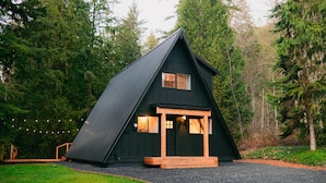 Eine schwarze Holzhütte mit einem dreieckigen Dach und einer Veranda sowie einer Auffahrt mit Kies.