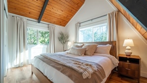 Ein Schlafzimmer mit schrägen Wänden, einem großen Bett mit Kopfteil, zwei Nachttischen mit Lampen und einem Fenster mit Vorhängen.