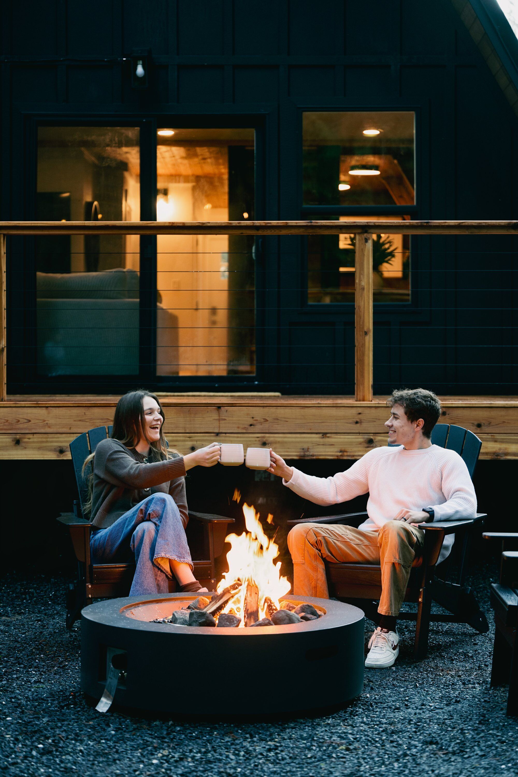 Deux personnes sont assises près d’un foyer extérieur, dans un cadre en plein air, avec une terrasse en bois et un bâtiment en arrière-plan.