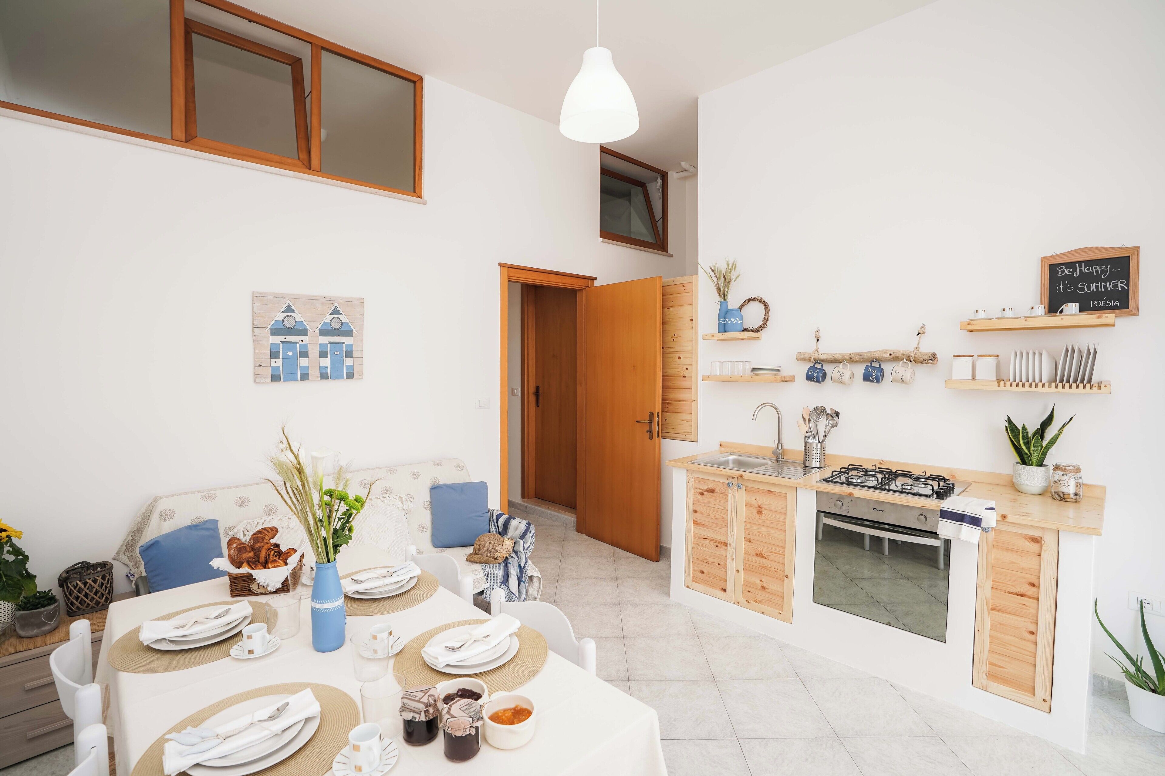 A compact kitchen with a gas stove, oven, and wooden cabinets. There is a dining area with a table set for two, a vase with flowers, and a wall-mounted shelf with kitchenware.