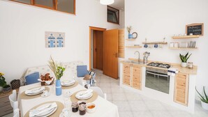 A compact kitchen with a gas stove, oven, and wooden cabinets. There is a dining area with a table set for two, a vase with flowers, and a wall-mounted shelf with kitchenware. - Poésia – Portuligno Apartment 50 mt from the most beautiful beaches in Salento! (melendugno)