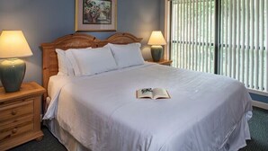 Una cama bien hecha con sábanas blancas, dos lámparas de noche, un cuadro en la pared y una ventana con persianas.