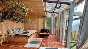 Een knus kamertje met een bed, een klein tafeltje en een bankje. Er is een groot raam met uitzicht op de omgeving.