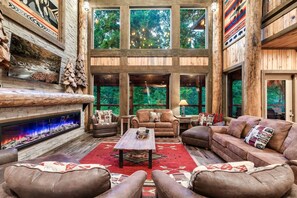 Ein geräumiges Wohnzimmer mit Kamin, Ledersofas und großen Fenstern mit Blick auf den Wald.