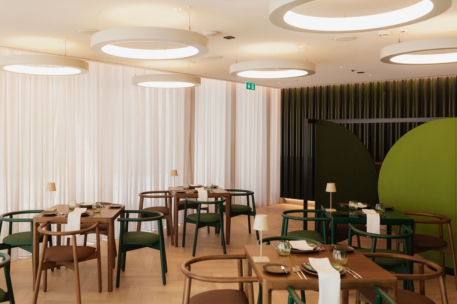Un espace repas moderne avec des tables rondes et des chaises vertes, agrémenté de luminaires circulaires au plafond et d’un mur d’accent vert.